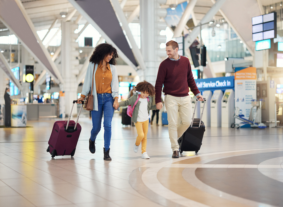 Family walking through an airport terminal with rolling luggage while their young daughter skips between them.