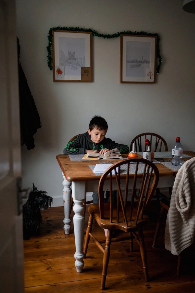 Child reading at a kitchen table in a lived-in family room, surrounded by chairs and everyday household objects.