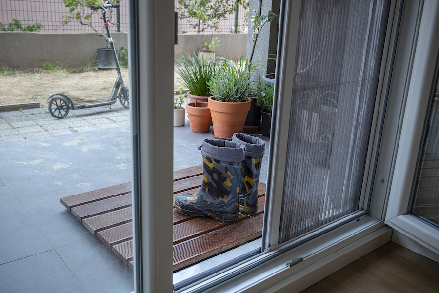 Child's rain boots beside a doorway, with potted plants and a scooter visible outside.